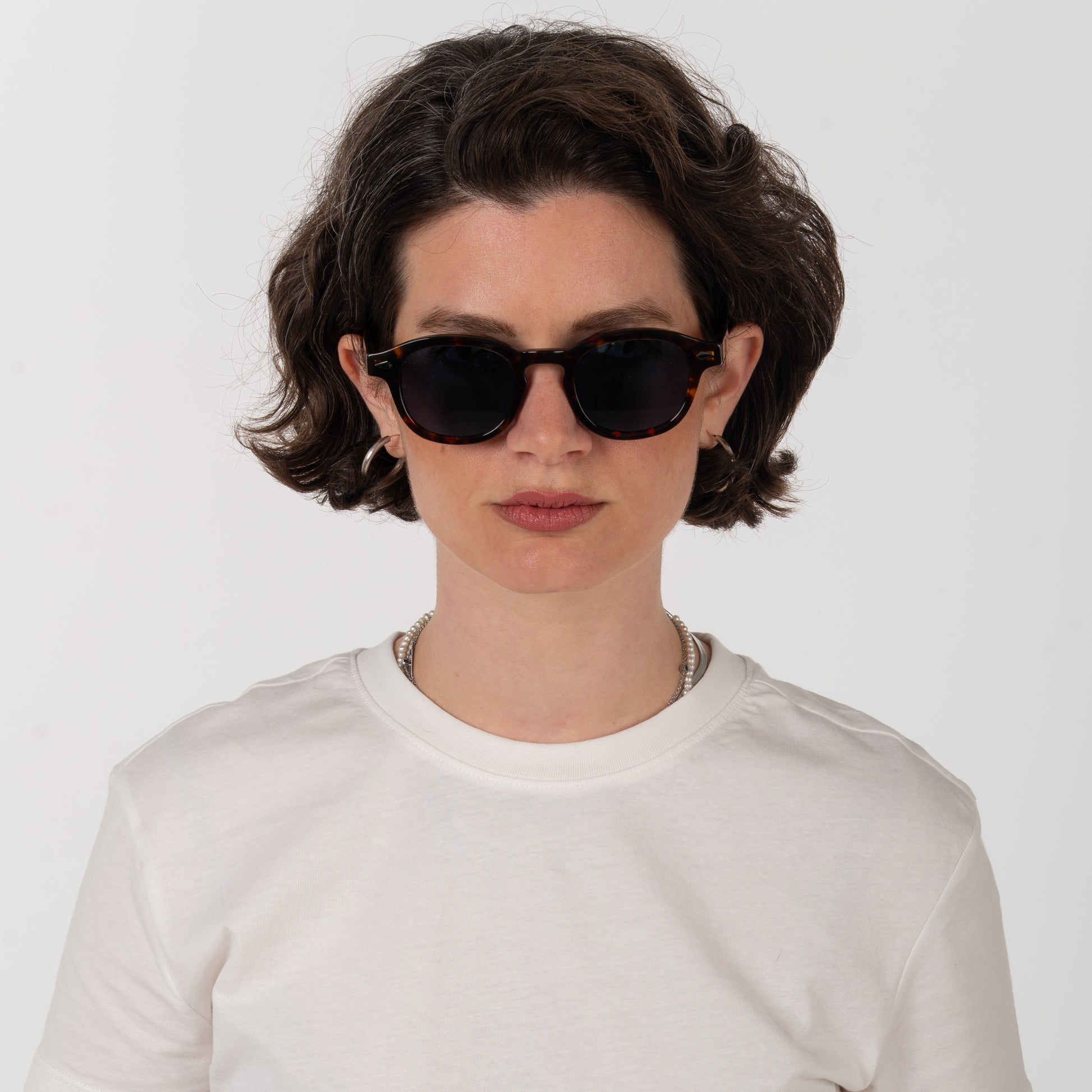 Person wearing dark sunglasses and a white shirt on a plain background
